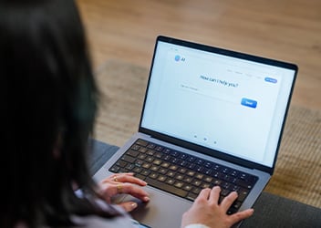 A person typing on a laptop with a web page open. The screen displays a text box and the question, "How can I help you?"